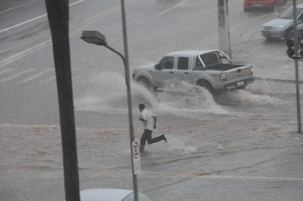 Rua alagada. Caminhonete passa jogando água para cima. Um pedrestre corre. | As chuvas intensas que ocorreram hoje no município, atingiram 70% do esperado para todo o mês de outubro.